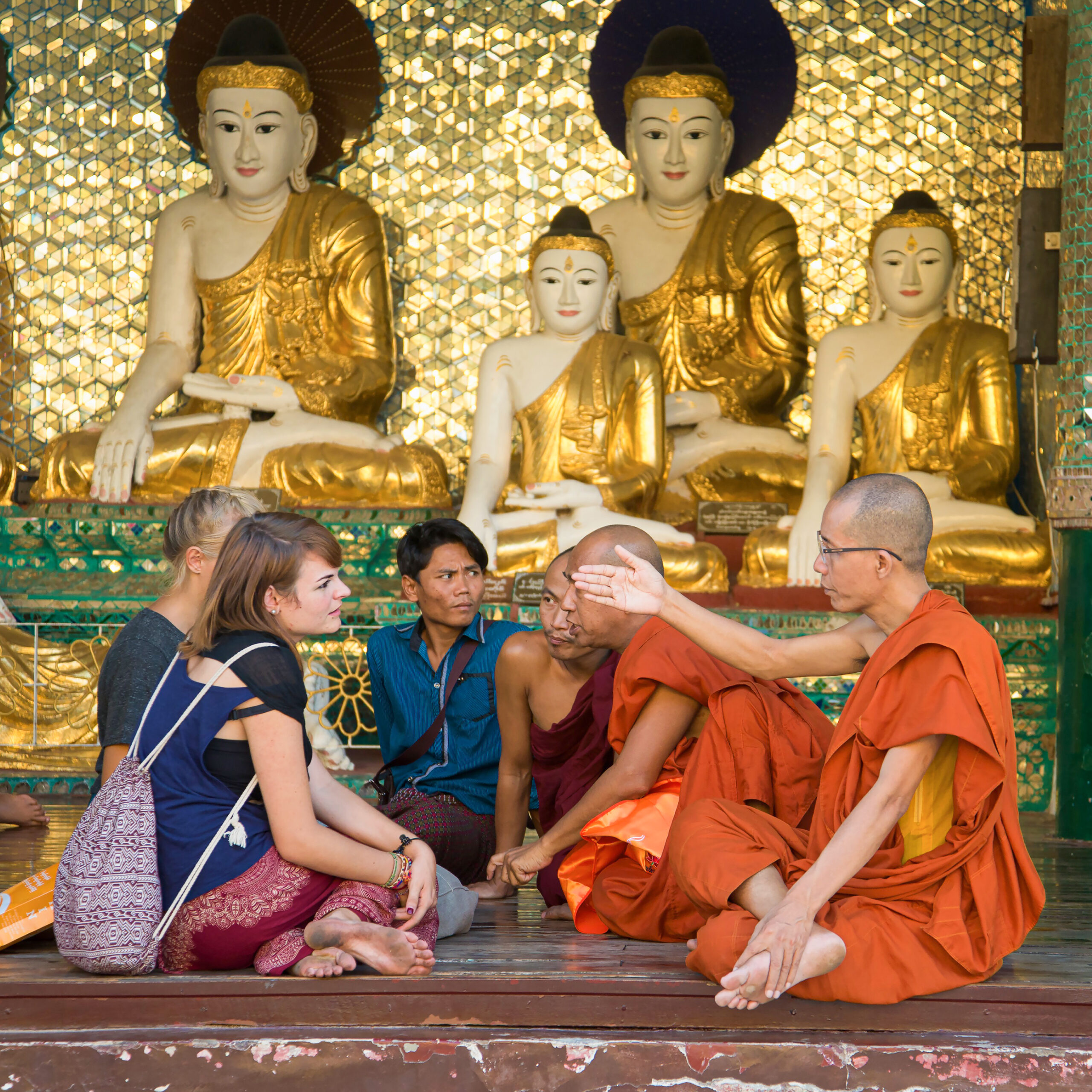 International travelers on an escorted group tour sit and converse with seated Buddhist monks.