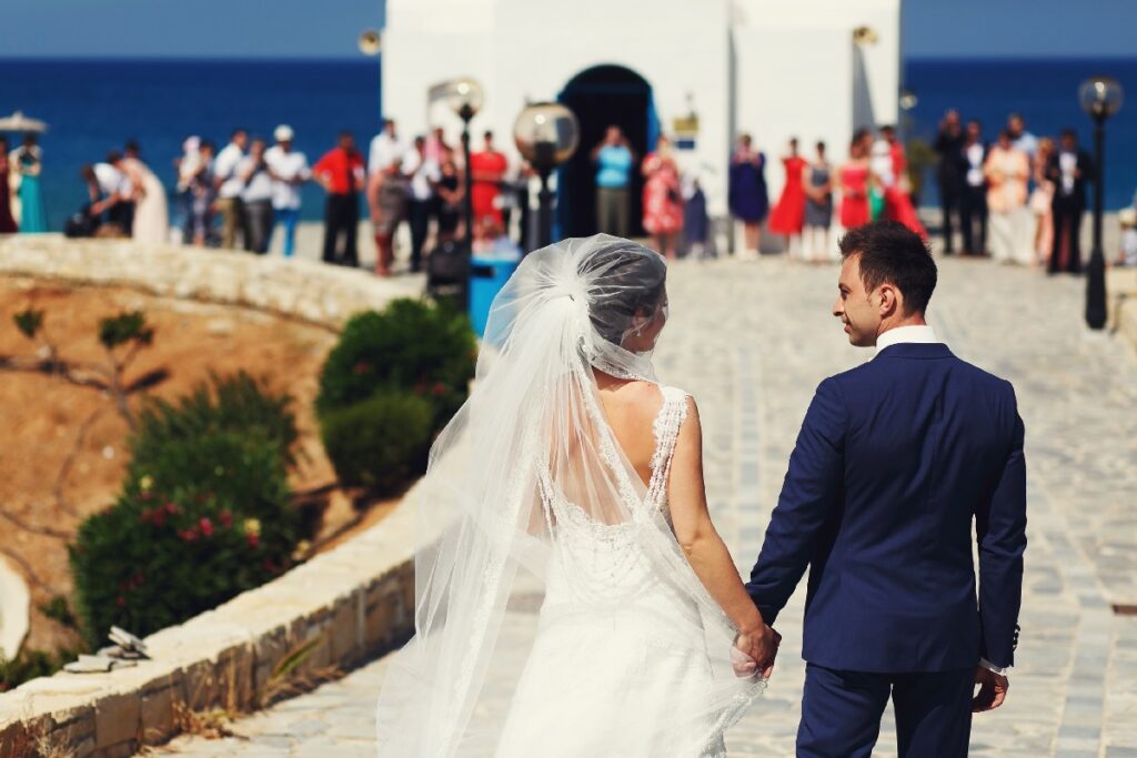 Bride in a lacy gown and groom in a blue suit walk hand in hand at a small jet-set destination wedding by the sea.