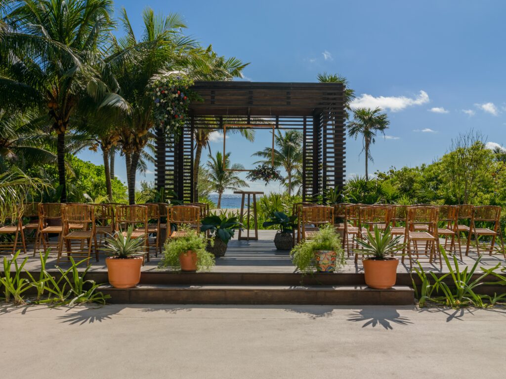 Raised beachfront gazebo with wedding chairs and decorative plants set up. Palm trees and ocean in the background.