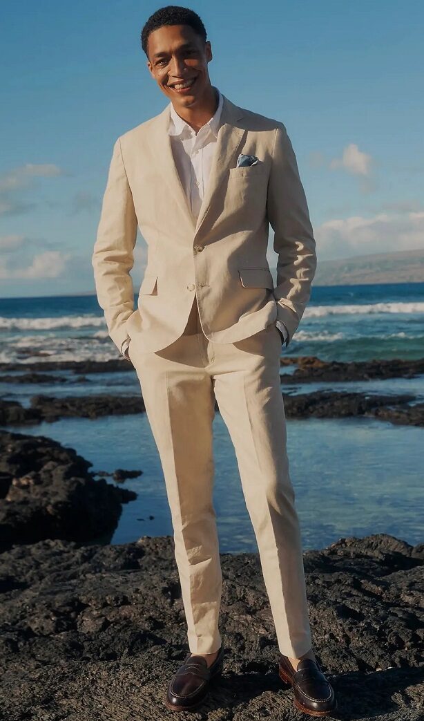 Elegantly dressed man in a beige suit, perfect for a destination wedding, stands on a beach.